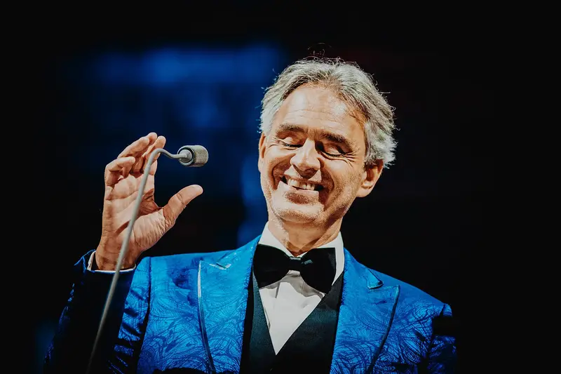 Andrea Bocelli smiling on stage with a microphone, in a blue jacket
