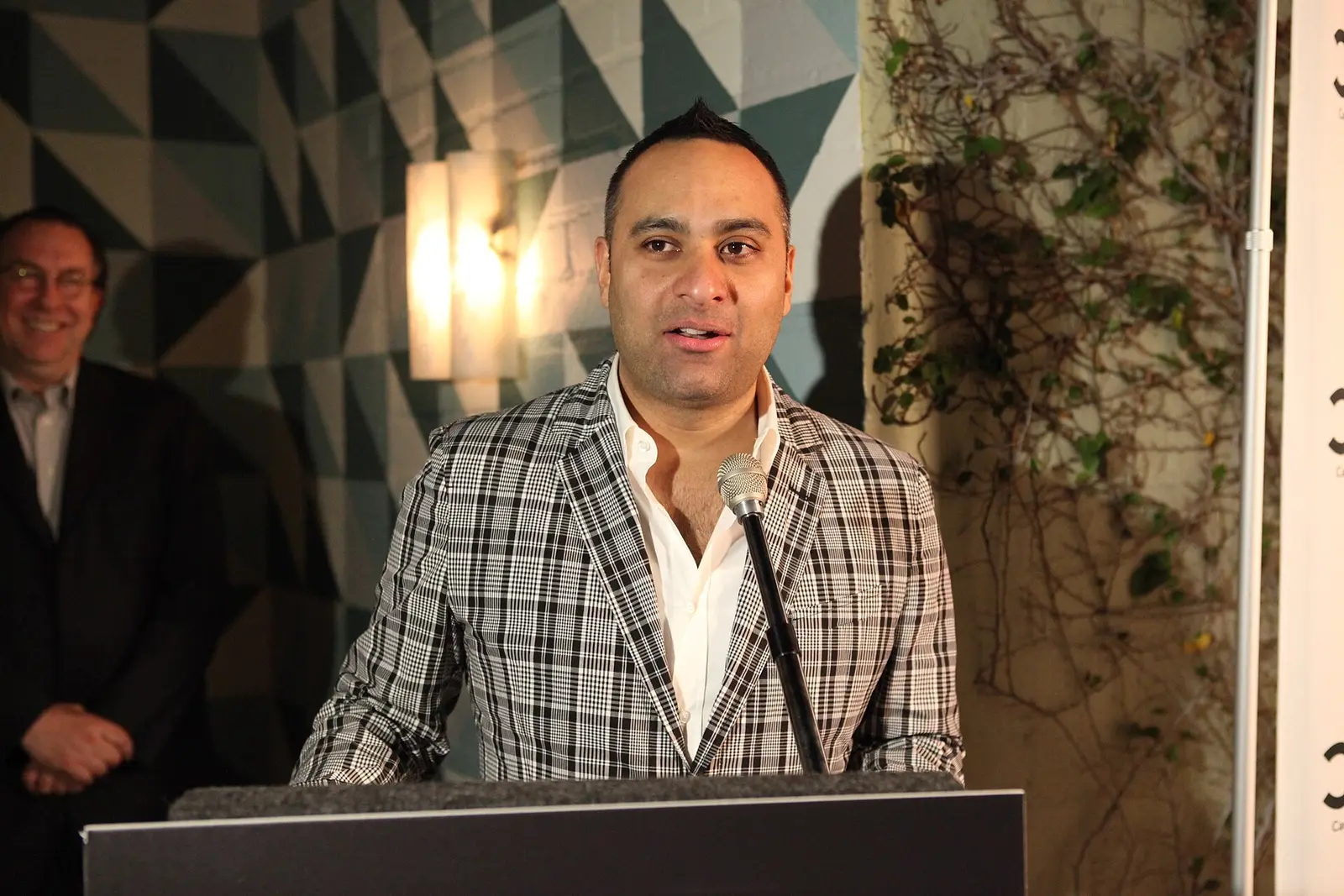 Russell Peters speaking at a podium in a checked jacket